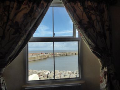 A window with floral curtains looking out to a harbor with a stone sea wall and water at Harbour Cottage in Watchet