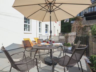 An outdoor patio with a round table and six chairs under a large umbrella with glasses and bottles on the table at Ferrystone Kingswear near Dartmouth