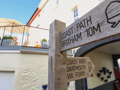 A wooden signpost indicating coast path directions to Brixham 10m and Dartmouth via ferry near a building with a balcony and umbrella at Ferrystone Kingswear near Dartmouth