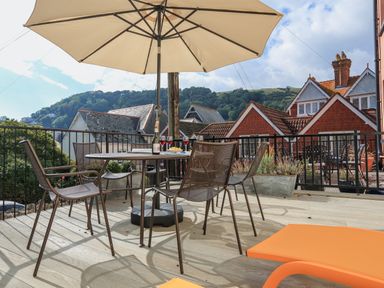 An outdoor patio with a round table and four chairs under a large beige umbrella with houses and hills in the background at Ferrystone Kingswear near Dartmouth