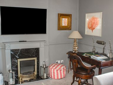 A corner room with a fireplace a wall-mounted TV a striped pouf a wooden desk with books two lamps and a wooden chair at Ferrystone in Kingswear near Dartmouth