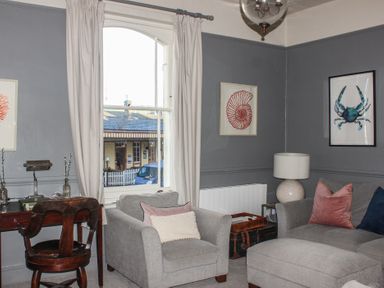 A living room with a gray armchair and sofa a wooden desk with a chair and framed artwork on the walls at Ferrystone in Kingswear near Dartmouth