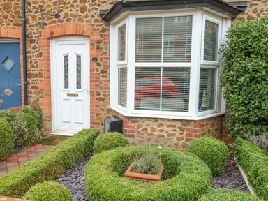 Front door and bay window of a house with brick and stone exterior and manicured bushes in garden at Hunny Cottage in Hunstanton