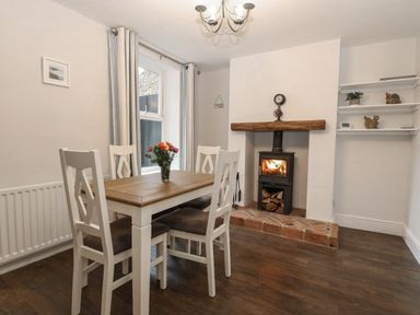 A dining area with a wooden table and four chairs a lit wood stove with firewood and shelves with ornaments at Hunny Cottage in Hunstanton