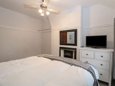 A bedroom with a bed, ceiling fan with lights, wall-mounted mirror, fireplace, white dresser with a television on top at Hunny Cottage in Hunstanton