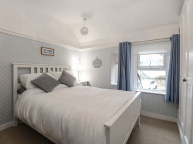 A bedroom with a white bed and grey pillows beside a window with blue curtains at Hunny Cottage in Hunstanton