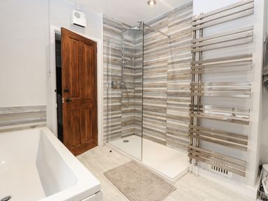A bathroom with a bathtub a wooden door a glass shower enclosure with striped tile wall and a chrome towel radiator at Hunny Cottage in Hunstanton