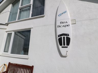 A white surfboard mounted on a white exterior wall next to two windows and a wooden bench at Sea Scape in Dinas Dinlle