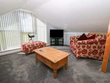 A sitting area with patterned armchairs a wooden coffee table a flat screen tv and large windows with vertical blinds at Sea Scape in Dinas Dinlle