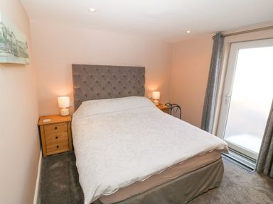 A bedroom with a bed a gray headboard two bedside tables with lamps and a door with gray curtains at Sea Scape in Dinas Dinlle