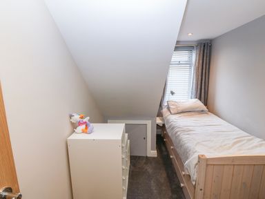 A small bedroom with a single bed a white dresser with a stuffed toy and a window with blinds and curtains at Sea Scape in Dinas Dinlle