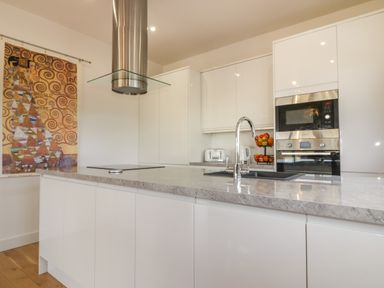 A modern kitchen with white cabinets a gray countertop a stovetop a stainless steel extractor fan a sink with a faucet a built in oven and microwave a toaster a kettle a fruit basket and a decorative tapestry on the wall at Woodhall in Cardinham