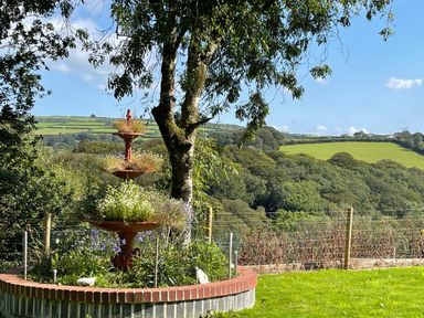A garden with a three-tier flower planter and a tree overlooking fields and hills at Woodhall in Cardinham