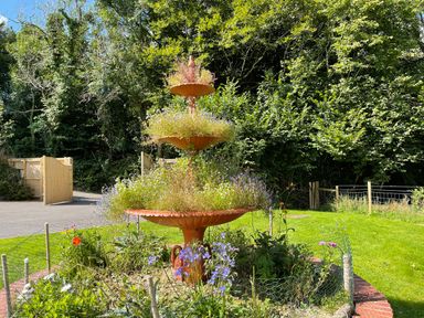 A tiered garden planter with plants and flowers in a fenced flower bed with trees and a wooden gate in the background at Woodhall in Cardinham