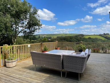 A wooden deck with a wicker sofa set and potted plants overlooking green hills and trees at Woodhall in Cardinham