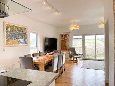A dining area with a wooden table and grey chairs a leather armchair and large windows showing an outdoor view at Woodhall in Cardinham