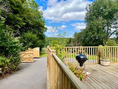 A wooden deck with a black barbecue grill and plants near a wooden fence and gate at Woodhall in Cardinham