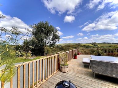 A wooden deck with railing, patio furniture, potted plants, overlooking trees and rolling hills under a blue sky at Woodhall in Cardinham