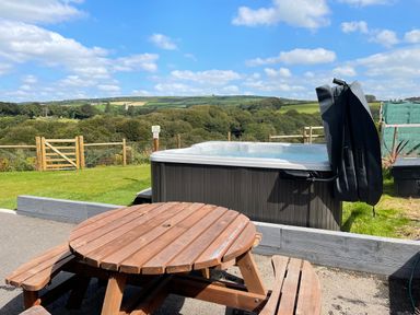 A wooden picnic table and benches near a hot tub with a countryside view at Woodhall in Cardinham