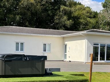A white single-story house with a flat roof and several windows with a hot tub and a lawn in the foreground at Woodhall in Cardinham