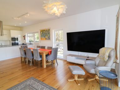 A dining area with a wooden table and gray chairs next to a kitchen and a living space with a large wall-mounted television and a leather armchair with an ottoman at Woodhall in Cardinham