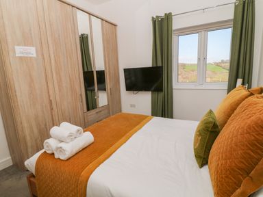 A bedroom with a bed made with orange and white bedding a set of rolled white towels on it a wall-mounted TV green curtains and a wooden wardrobe with mirrors at Woodhall in Cardinham