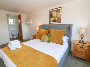 A bedroom with a bed with yellow pillows and blanket two wooden nightstands with lamps an open door showing bathroom with a toilet and a white clothes rack at Woodhall in Cardinham