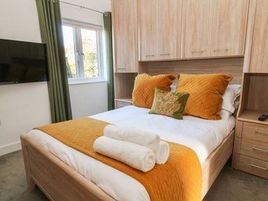 A bedroom with a bed with orange pillows and blanket two white rolled towels on the bed green curtains and a wall mounted tv at Woodhall in Cardinham