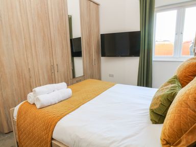 A bedroom with a bed featuring orange and green pillows and blanket with rolled white towels on top wooden wardrobes against one wall and a wall-mounted television near a window with green curtains at Woodhall in Cardinham