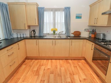 A kitchen with light wood cabinets a double sink under a window with blue curtains and wooden floor at 2 Chandlers Yard in Burry Port