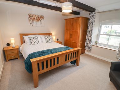 A bedroom with a wooden bed frame bedside tables a wooden wardrobe a window with curtains and a ceiling light at The Cottage in Tetbury