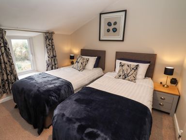 A bedroom with two single beds with dark blue throws and patterned cushions a bedside table with a lamp and a window with curtains at The Cottage in Tetbury
