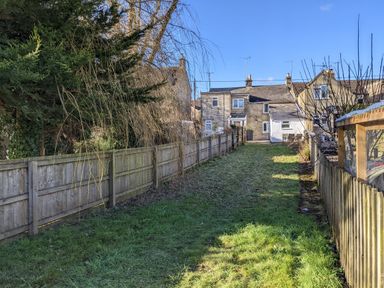 A grassy backyard area fenced with wooden panels leading to stone houses under a blue sky