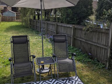 A garden with two black lounge chairs under an umbrella beside a small table with a drink at The Cottage in Tetbury