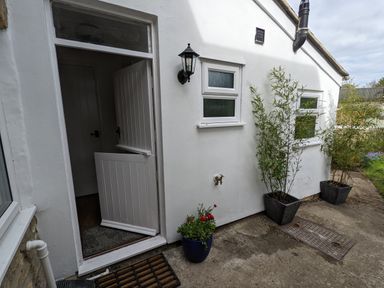 An open white half door on a white exterior wall with two small windows two potted plants and a garden tap at The Cottage in Tetbury