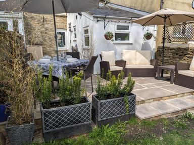 An outdoor patio with a dining table set with dishes and glasses a seating area with cushioned chairs and umbrellas and plants in planters at The Cottage in Tetbury
