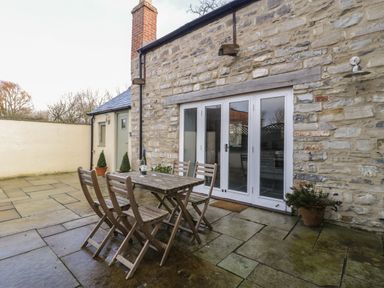 An outdoor patio area with a wooden table and four chairs next to stone wall with white doors and potted plants at Magnolia Barn in Mark