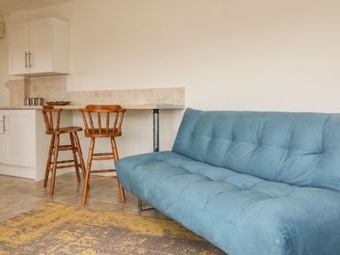 A kitchen area with white cabinets and a tiled backsplash with two wooden bar stools and a blue sofa with a patterned rug in front at The Stables in Crowcombe