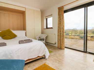 A bedroom with a bed white linens a green pillow towels on bed built in wardrobe a wicker chair tiled floor and sliding glass door overlooking a patio and countryside at The Stables in Crowcombe