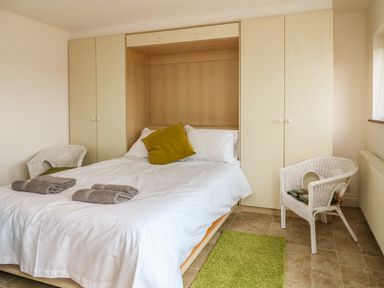 A bedroom with a double bed with white bedding and a green pillow two white wicker chairs wardrobes and a green rug at The Stables in Crowcombe