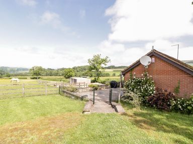 A fenced garden with a patio and flowers next to a brick building and a white horse in a field at The Stables in Crowcombe