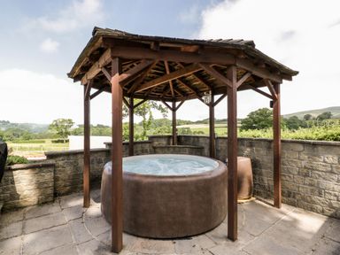 An outdoor hot tub under a wooden gazebo with stone walls and countryside views at The Stables in Crowcombe