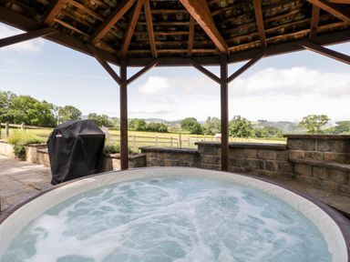 A hot tub under a wooden gazebo with a barbecue and countryside view at The Stables in Crowcombe