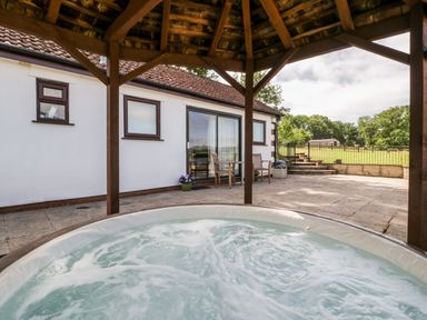 A patio with a hot tub under a wooden gazebo and a table with two chairs near a white building at The Stables in Crowcombe