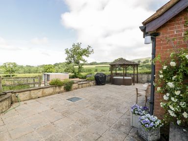 An outdoor patio area with a wooden gazebo over a hot tub a barbecue grill potted flowers and countryside views at The Stables in Crowcombe