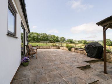 An outdoor patio with stone tiles a covered barbecue a wooden chair and table a potted plant and a wooden fence with trees in the background at The Stables in Crowcombe