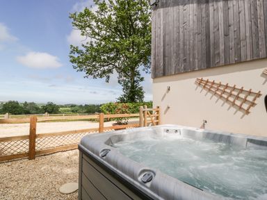An outdoor hot tub next to a building with wood paneling and a wooden fence with a tree and fields in the background at The Outlook in Crowcombe
