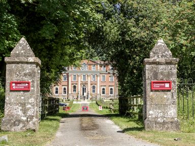A driveway between two stone pillars leading to a large brick building surrounded by trees at Crowcombe Court in Crowcombe