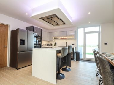 A modern kitchen with a refrigerator island with two bar stools and a dining table with chairs by a glass door at Valley View in Mullion