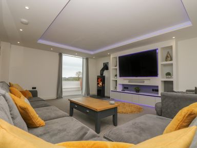 A living room with a gray sofa and yellow cushions a wooden coffee table a wall-mounted television and a wood-burning stove at Valley View in Mullion